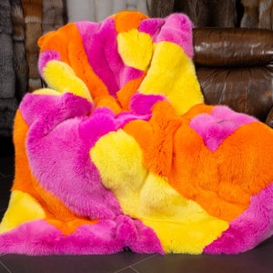 Può includere: Una vivace coperta in finta pelliccia, drappeggiata su una poltrona in pelle marrone. La coperta presenta ampie macchie di rosa acceso, giallo e arancione, creando un design audace. La texture morbida è evidente.