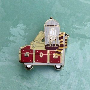 May include: Enamel pin showing a red and gold trunk on wheels, with luggage and a cage holding a white owl. A striped scarf is draped over the cage. The pin has gold detailing.