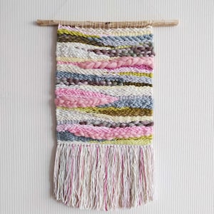Peut inclure: Une tenture murale tissée avec une tringle en bois. Le textile présente des rayures horizontales dans les tons de rose, gris, jaune et blanc, avec un bas texturé et frangé. La pièce est suspendue par une fine ficelle.