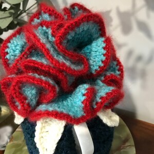 May include: A crocheted mug cozy in blue, red, and white. The cozy is shaped like a flower with ruffled edges.