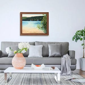 May include: A framed painting depicting a lake scene with trees and a sandy shore. The painting is hanging on a white wall above a grey sofa with cushions and a throw blanket.