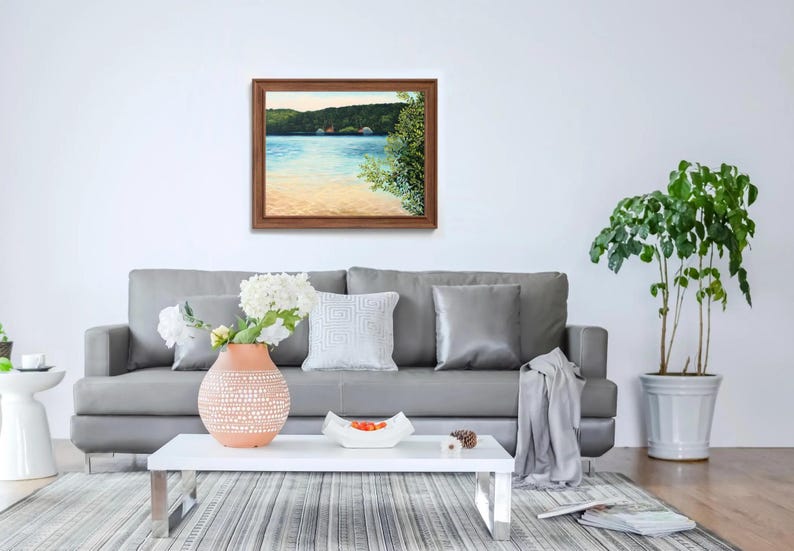 May include: A framed painting depicting a lake scene with trees and a sandy shore. The painting is hanging on a white wall above a grey sofa with cushions and a throw blanket.