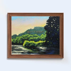 May include: A framed oil painting depicting a tranquil river scene. Lush green trees line the riverbank, with a dark green tree in the foreground. The sky is a soft blue with hints of pink and orange, suggesting a sunset or sunrise.
