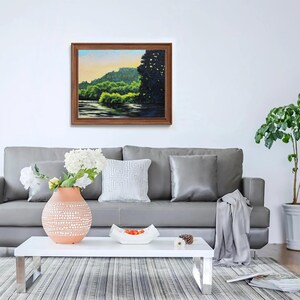 May include: A gray sofa with a throw blanket and two pillows sits in a living room with a white coffee table and a potted plant. A framed painting of a green landscape hangs on the wall above the sofa.