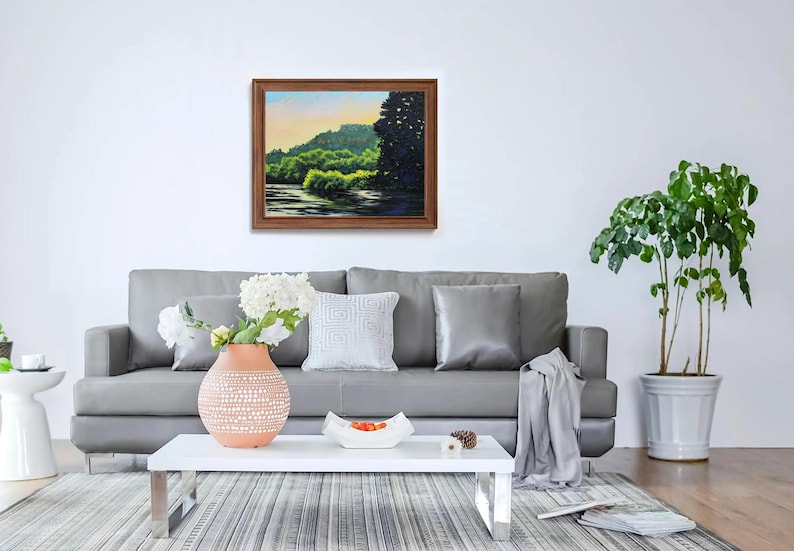 May include: A gray sofa with a throw blanket and two pillows sits in a living room with a white coffee table and a potted plant. A framed painting of a green landscape hangs on the wall above the sofa.