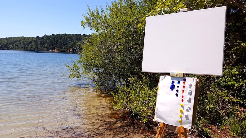 May include: A white canvas on an easel with a paint palette with blue, red, yellow, and white paint. The easel is set up on a sandy beach with a lake in the background.