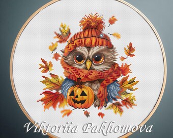 May include: A cross-stitch embroidery project in a round wooden hoop featuring a brown owl wearing an orange knit hat and scarf, holding a small orange jack-o'-lantern pumpkin, surrounded by colorful autumn leaves with the text Viktoriia Pakhomova.