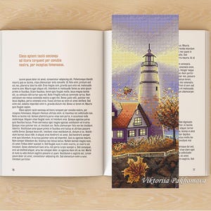 Puede incluir: Un marcapáginas hecho a mano con una escena de faro con una casa, árboles y hojas de otoño. El marcapáginas está colocado sobre un libro abierto con texto en latín. El nombre del artista, Viktoriia Pakhomova, es visible en la parte inferior.