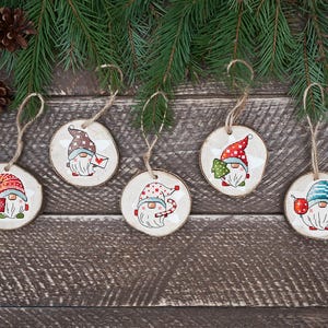 Puede incluir: Cinco adornos de madera con forma de círculo con un diseño de gnomo. Cada gnomo lleva un sombrero de un color diferente y tiene un diseño diferente en su ropa. Los adornos están colgados de un cordel.