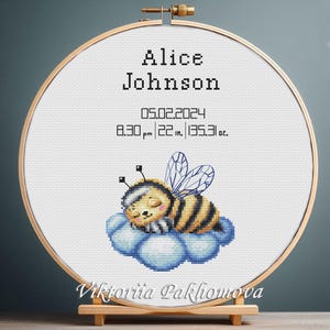 May include: A white embroidery hoop with a cross-stitch design of a sleeping bee on a cloud. The text "Alice Johnson 05.02.2024 8:30 pm | 22 in. | 135.31 oz." is embroidered below the bee. The text "Viktoria Pakhomova" is embroidered at the bottom of the hoop.