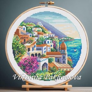 Peut inclure: Une broderie au point de croix d'une ville côtière italienne, encadrée dans un cercle en bois. La scène représente des bâtiments colorés, une végétation luxuriante et une église avec un dôme, sur fond de mer et de ciel bleus. Le texte "Viktoriia Pakhomova" est en bas.