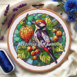 Könnte beinhalten: Kreuzstich-Stickerei in einem Holzreifen mit einem bunten Vogel, Beeren und Blättern. Das Design enthält den Text "Viktoriia Pakhomova". Der Vogel hat rote, orangefarbene, violette und schwarze Federn. Der Hintergrund ist ein Glasmalerei-Effekt.