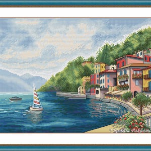 May include: Cross-stitch depiction of a coastal town with vibrant buildings, a sailboat, and a mountain backdrop. The artwork features a blue sea, green hills, and a cloudy sky. The image is framed in a blue border.