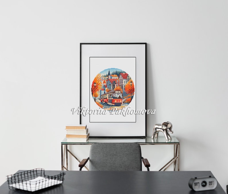 May include: A framed watercolor print depicting a European town scene with a red trolley car. The artwork is in a circular format, framed in black, and features autumn colors. The artist's name, Viktoriia Pakhomova, is printed in cursive.