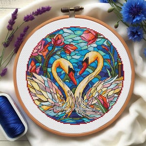 May include: Cross-stitch embroidery of two swans forming a heart shape, set against a stained-glass style background. The swans have yellow necks and red beaks. The design is framed in a wooden hoop, with blue thread and flowers in the background.