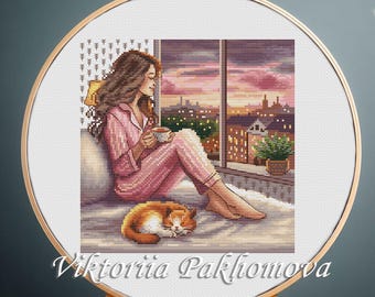 May include: Cross-stitch embroidery in a wooden hoop depicts a person in pink pajamas, holding a cup, looking out a window at a sunset over a city. A sleeping orange cat rests on the bed. The text "Viktoriia Pakhomova" is at the bottom.