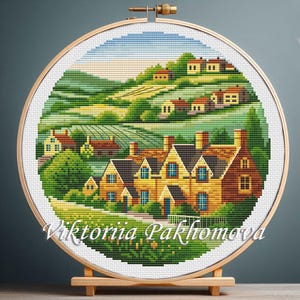May include: A cross-stitch landscape scene of a village with houses, trees, and rolling hills. The artwork is displayed in a round wooden embroidery hoop. The text "Viktoriia Pakhomova" is at the bottom of the image.
