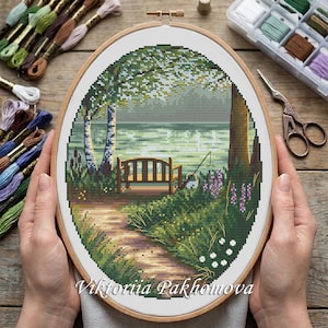 May include: An oval embroidery hoop containing a cross-stitch pattern of a park scene with a wooden bench, a dirt path, birch trees, and a lake. The design features green, brown, and white threads. The text Viktoriia Pakhomova is at the bottom.