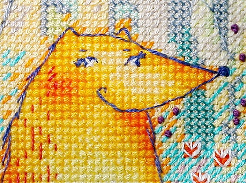 Op de afbeelding: Een close-up van een gele vos met blauwe ogen en een glimlach, geborduurd op een witte stof met een kleurrijk bloemenpatroon.