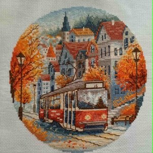 May include: Cross-stitch embroidery of a European city scene with a red tram, buildings, and autumn foliage. The artwork is circular, with a detailed depiction of a street scene, including streetlights and a bench. The color palette includes reds, oranges, blues, and greens.