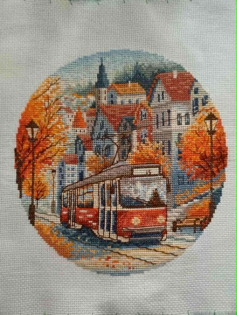 May include: Cross-stitch embroidery of a European city scene with a red tram, buildings, and autumn foliage. The artwork is circular, with a detailed depiction of a street scene, including streetlights and a bench. The color palette includes reds, oranges, blues, and greens.