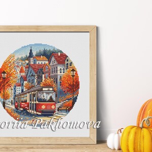 May include: Framed cross-stitch art depicting a red trolley traveling through a European town in autumn. The artwork features orange and yellow trees, red-roofed buildings, and a blue sky. The frame is light brown, and the artist's name is visible.