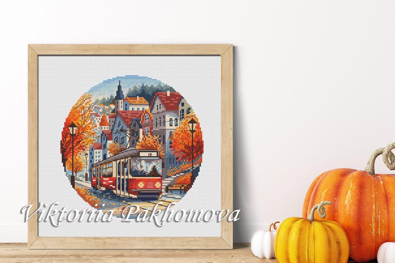 May include: Framed cross-stitch art depicting a red trolley traveling through a European town in autumn. The artwork features orange and yellow trees, red-roofed buildings, and a blue sky. The frame is light brown, and the artist's name is visible.