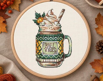 May include: A cross-stitch pattern in a wooden embroidery hoop featuring a mason jar mug filled with a beverage topped with whipped cream and a straw. The mug has a green textured cozy with the text FALL VIBES printed on a central label.