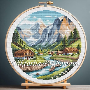 Könnte beinhalten: Eine Kreuzstich-Landschaftsszene mit einem Gebirge, einem Fluss und Häusern, präsentiert in einem hölzernen Stickrahmen. Das Kunstwerk zeigt eine detaillierte Darstellung eines Tals mit grünen Bäumen, blauem Wasser und braunen Häusern. Der Text "Viktoriia Pakhomova" befindet sich unten.