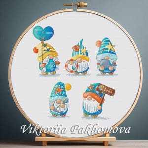May include: Cross-stitch embroidery of five beach gnomes in a wooden hoop. Each gnome wears a blue and yellow hat and holds beach-themed items like a balloon, life preserver, and a sign that says "Beach". The artist's name, Viktoriia Pakhomova, is at the bottom.