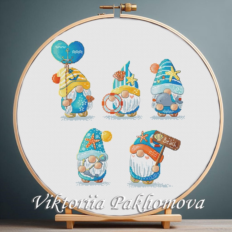 May include: Cross-stitch embroidery of five beach gnomes in a wooden hoop. Each gnome wears a blue and yellow hat and holds beach-themed items like a balloon, life preserver, and a sign that says "Beach". The artist's name, Viktoriia Pakhomova, is at the bottom.