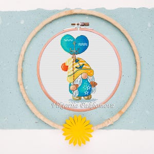 May include: Cross-stitch embroidery of a gnome holding a heart-shaped balloon with an anchor design. The gnome wears a blue outfit with star details, a yellow hat, and holds a seashell. The embroidery is in a wooden hoop, with a yellow flower below.