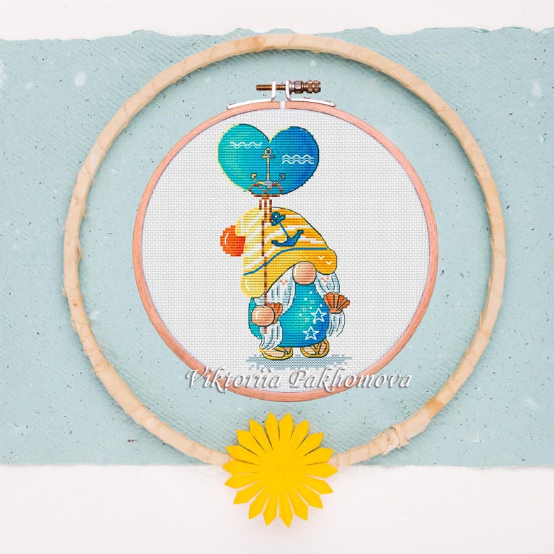 May include: Cross-stitch embroidery of a gnome holding a heart-shaped balloon with an anchor design. The gnome wears a blue outfit with star details, a yellow hat, and holds a seashell. The embroidery is in a wooden hoop, with a yellow flower below.