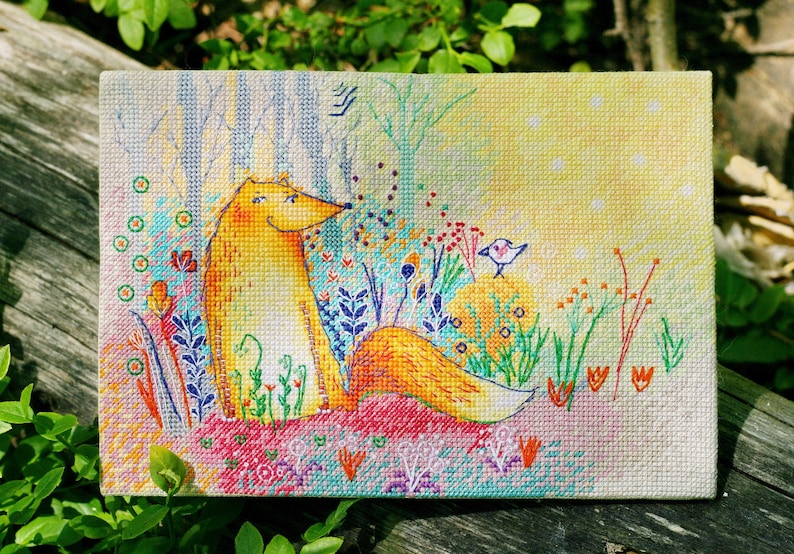 Op de afbeelding: Een borduurwerk met kruissteek van een gele vos die in een kleurrijke weide zit met een kleine blauwe vogel die op een tak zit. De achtergrond is lichtgeel met witte stippen.