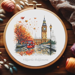 Op de afbeelding: Kruissteekborduurwerk van de Big Ben en een rode dubbeldekkerbus in Londen in de herfst. Het kunstwerk is ingelijst in een houten hoepel en toont vallende bladeren en de naam van de kunstenaar, Viktoriia Pakhomova.