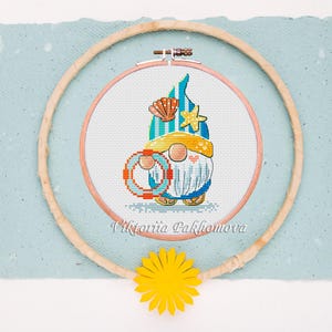 May include: Cross-stitch embroidery of a beach gnome wearing a yellow hat and holding a life preserver. The gnome is inside a wooden embroidery hoop, with a yellow paper flower at the bottom. The text "Viktoriia Pakhomova" is at the bottom.