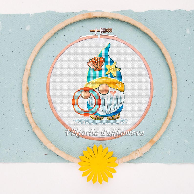 May include: Cross-stitch embroidery of a beach gnome wearing a yellow hat and holding a life preserver. The gnome is inside a wooden embroidery hoop, with a yellow paper flower at the bottom. The text "Viktoriia Pakhomova" is at the bottom.