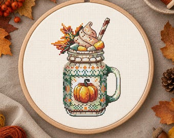 May include: A cross-stitch pattern in a wooden embroidery hoop featuring a mason jar mug filled with a pumpkin-themed beverage, topped with whipped cream and a cinnamon stick, with the text Viktoriia Pakhomova at the bottom.