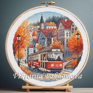 May include: A cross-stitch embroidery of a city scene with a red tram, buildings, and autumn trees. The artwork is displayed in a wooden hoop and mounted on a wooden stand. The text "Viktoriia Pakhomova" is at the bottom.