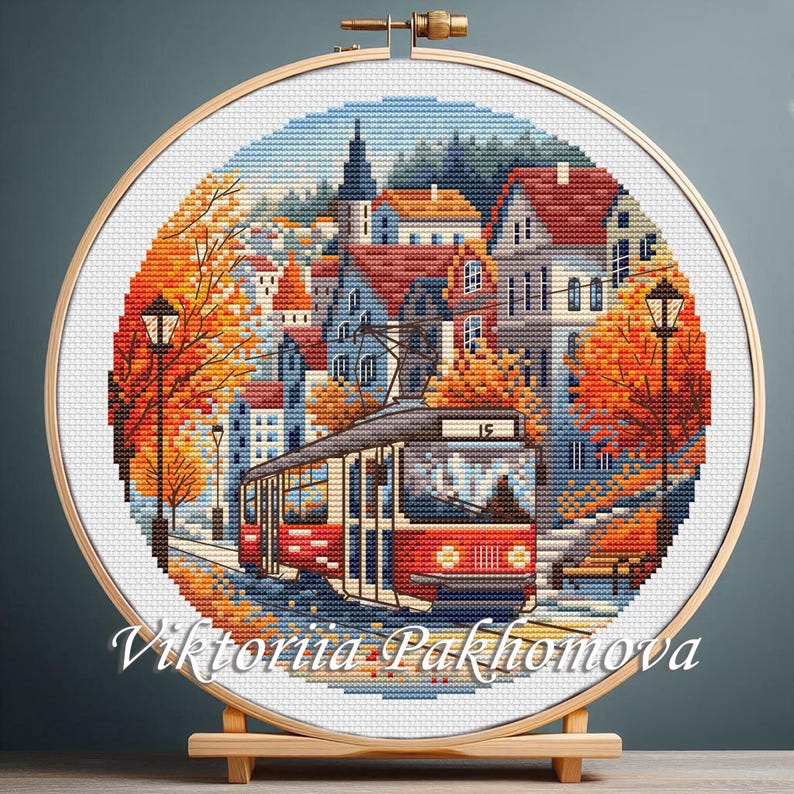 May include: A cross-stitch embroidery of a city scene with a red tram, buildings, and autumn trees. The artwork is displayed in a wooden hoop and mounted on a wooden stand. The text "Viktoriia Pakhomova" is at the bottom.