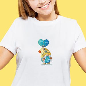 May include: White t-shirt featuring a colorful gnome design. The gnome is holding a blue heart-shaped balloon with a nautical theme. The gnome wears a yellow hat and blue outfit with star and seashell details.