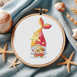 May include: Cross-stitch embroidery in a wooden hoop features a gnome with a mermaid tail and starfish. The gnome's hat is red with gold stars, and the tail is orange and red. The design is surrounded by seashells and starfish.
