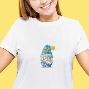 May include: White t-shirt featuring a colorful embroidered gnome design. The gnome wears a blue hat with a starfish and holds a pair of binoculars. A yellow ball is attached to the hat. The gnome is wearing a white beard and a blue and white outfit.