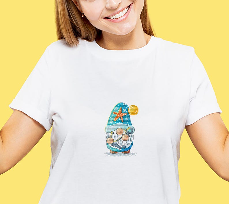 May include: White t-shirt featuring a colorful embroidered gnome design. The gnome wears a blue hat with a starfish and holds a pair of binoculars. A yellow ball is attached to the hat. The gnome is wearing a white beard and a blue and white outfit.