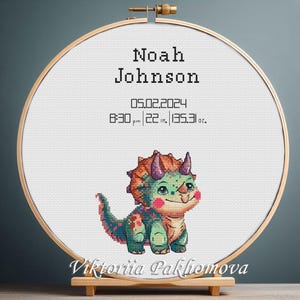 Op de afbeelding: Kruissteekkunst in een houten hoepel, met een vriendelijke dinosaurus in groen en oranje. De tekst luidt "Noah Johnson" met geboortegegevens: 55,88 cm, 3830,6 g en de datum 05.02.2024. De hoepel wordt op een houten standaard gepresenteerd.