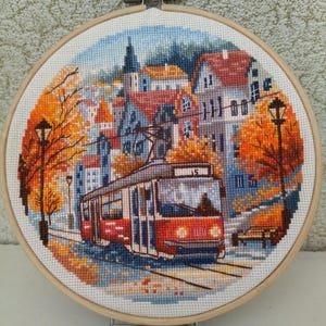 May include: Cross-stitch embroidery of a European town scene with a red tram, buildings, and autumn trees. The artwork is framed in a wooden hoop, showcasing a detailed landscape with vibrant colors and intricate stitching.