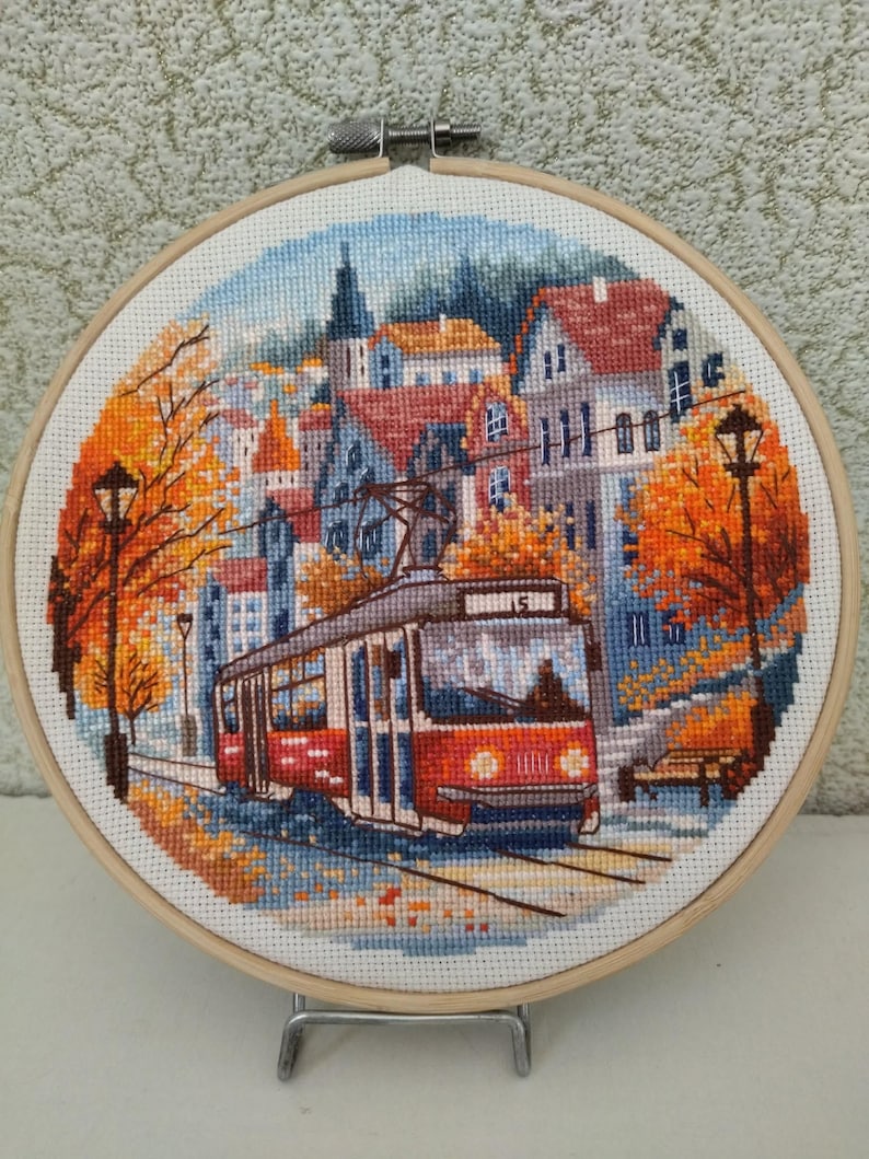 May include: Cross-stitch embroidery of a European town scene with a red tram, buildings, and autumn trees. The artwork is framed in a wooden hoop, showcasing a detailed landscape with vibrant colors and intricate stitching.