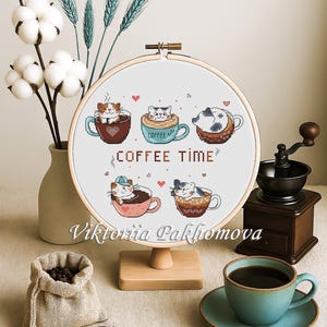 May include: A cross-stitch embroidery hoop with a design of cats in coffee cups and the words "COFFEE TIME". The embroidery is displayed on a wooden stand, with a cup of coffee, coffee beans, and a coffee grinder in the background.
