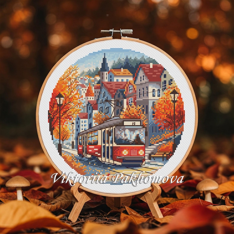 May include: A cross-stitch embroidery of a city scene with a red tram, buildings, and autumn trees. The embroidery is in a wooden hoop, displayed on a wooden easel. The artist's name, Viktoriia Pakhomova, is written at the bottom.
