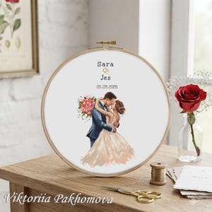 Peut inclure: Un tambour à broder circulaire en bois contenant un motif au point de croix représentant un couple qui s'enlace. Le texte indique Sara, Jes et 05.09.2026, avec deux anneaux dorés. Le couple porte un costume bleu et une robe de mariée blanche.